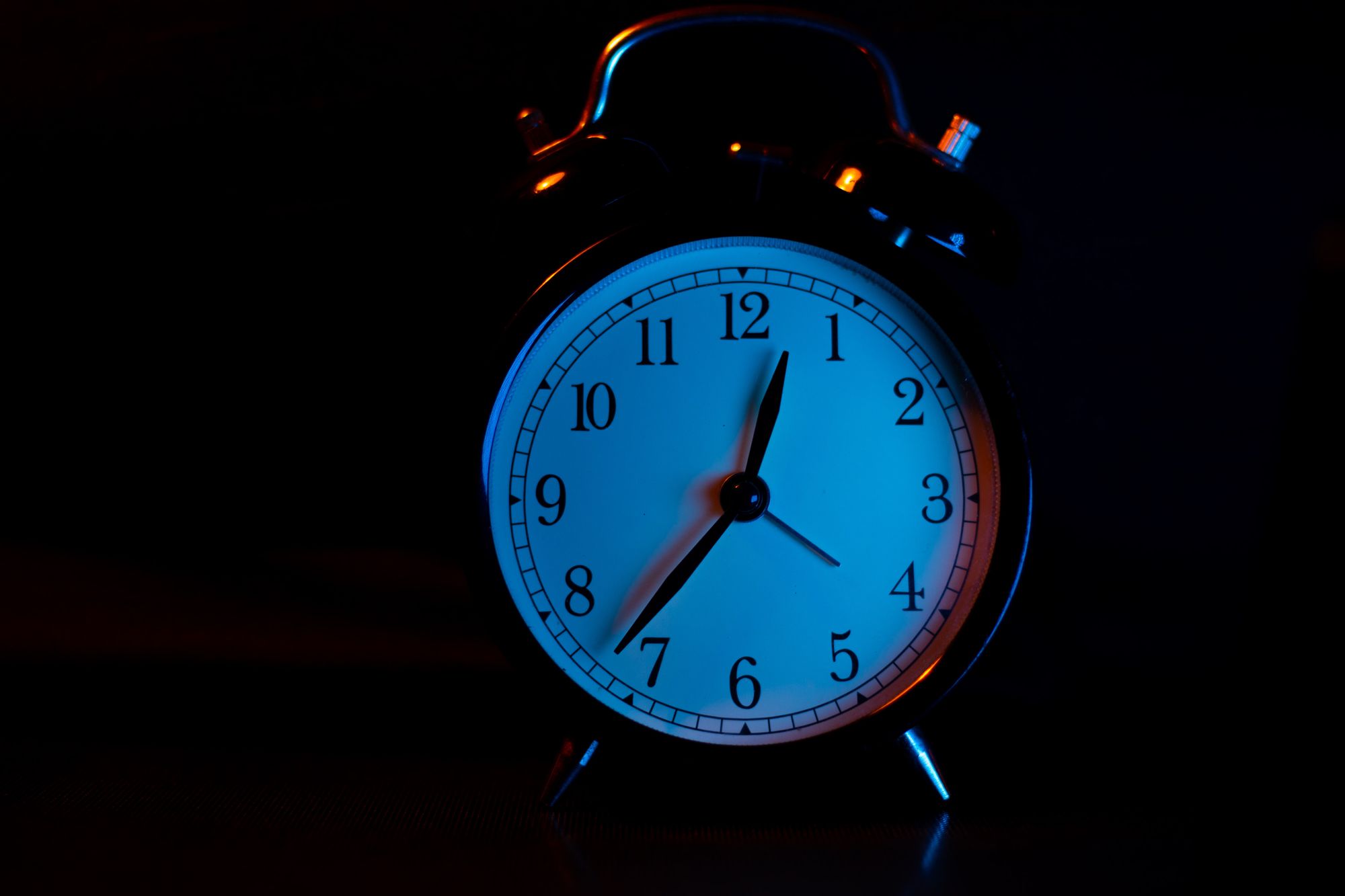 Dark background, photo of classic belle top alarm clock, white face, analog, 2 hands clock face, numbers, 