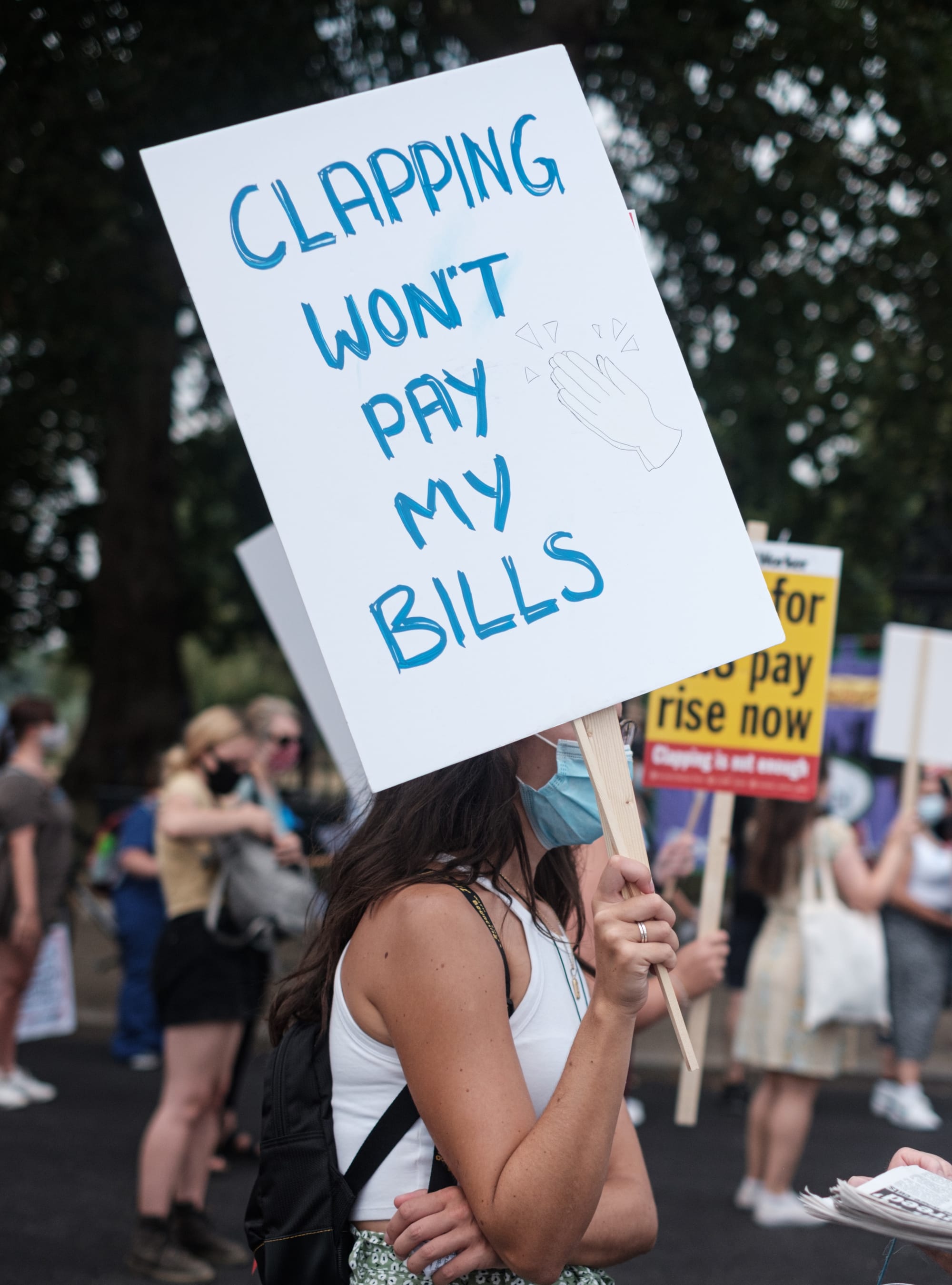 Protester in a surgical mask outside surrounded by people, holding a sign "Clapping won't pay my bills."