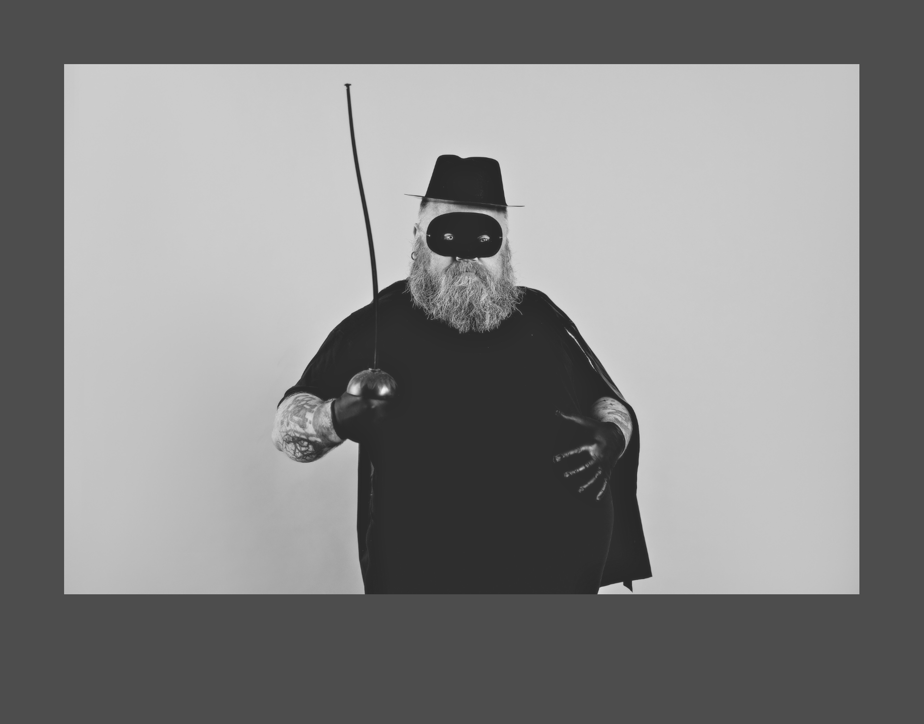 Bearded man in kids black mask and black gloves, came, and hat with fencing sword and big round belly.