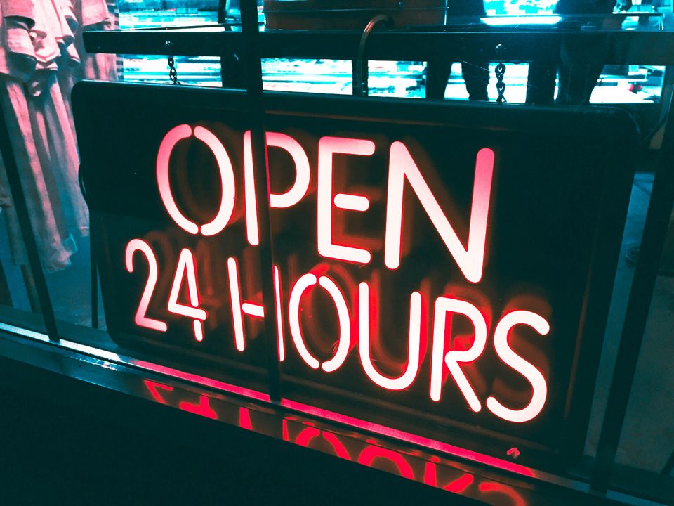 Neon sign in pink "Open 24 hours" on a street level gated window.